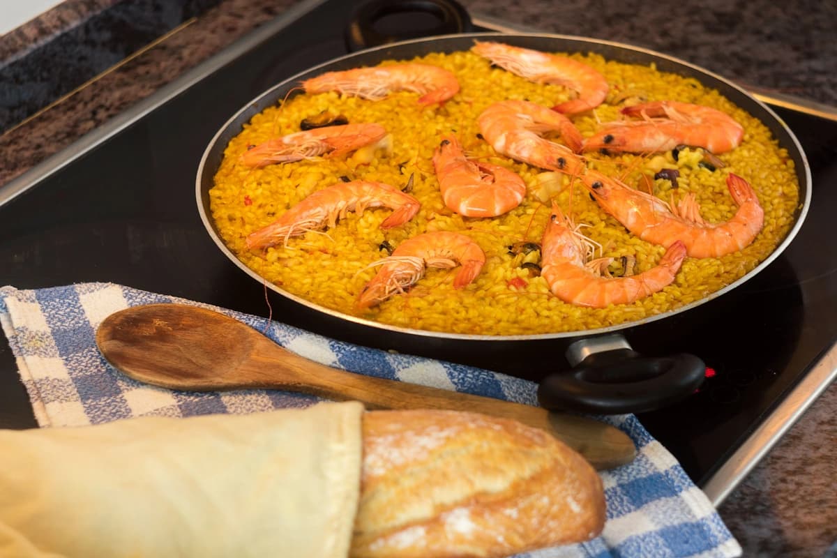 A pan of paella with shrimp on the kitchen table, with bread nearby.